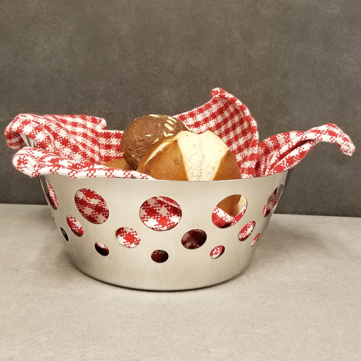A stainless steel basket with round cutout patterns holds two pretzel rolls. Lined with a red and white checkered cloth, the dishwasher safe basket sits on a plain gray surface, creating a simple, modern look.
