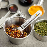 A stainless steel measuring spoon labeled 1 tsp rests in a cup filled with crushed red pepper flakes. Nearby, metal cups hold dried herbs and bright yellow turmeric powder, plus assorted measuring spoons—all on a gray countertop.