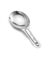 A shiny stainless steel measuring spoon with a short handle is shown on a white background. The handle is stamped with measurement markings: 1 TBSP and 15 mL. A small hole at the end allows for hanging, and it is dishwasher safe.
