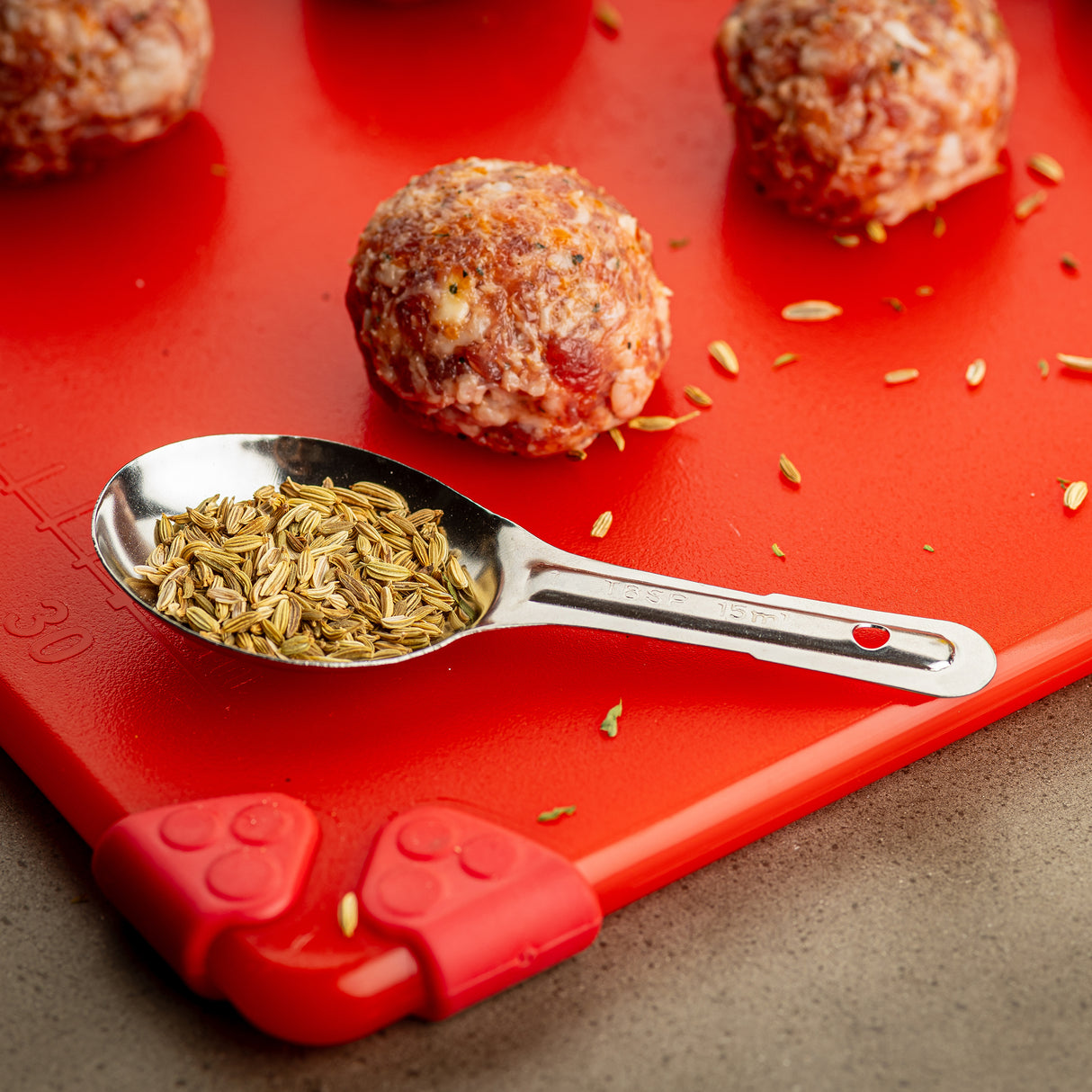 A stainless steel measuring spoon filled with fennel seeds rests on a bright red cutting board. Nearby, raw meatballs with visible spices sit among scattered fennel seeds. The board features red silicone corners and sits on a gray countertop.