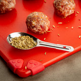 A stainless steel measuring spoon filled with fennel seeds rests on a bright red cutting board. Nearby, raw meatballs with visible spices sit among scattered fennel seeds. The board features red silicone corners and sits on a gray countertop.