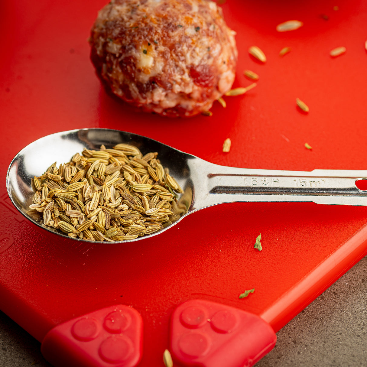 A stainless steel measuring spoon filled with fennel seeds rests on a red cutting board. Nearby, a raw meatball and scattered seeds are visible. The bright light highlights the textures of the seeds, cutting board, and shiny, dishwasher safe spoon.