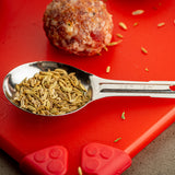 A stainless steel measuring spoon filled with fennel seeds rests on a red cutting board. Nearby, a raw meatball and scattered seeds are visible. The bright light highlights the textures of the seeds, cutting board, and shiny, dishwasher safe spoon.