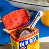 A stainless steel measuring spoon rests inside an open Old Bay Seasoning container. The red lid is flipped open, showing the brownish-orange spice inside. In the background are lemon wedges and seafood on a blue cutting board.