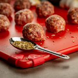 A stainless steel measuring spoon filled with fennel seeds rests on a red cutting board. Behind it, several raw meatballs are arranged in rows, ready to be cooked. The blurred background highlights the fresh ingredients up close.