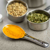 A stainless steel measuring spoon filled with bright yellow ground turmeric sits on a gray surface. Behind it, two metal measuring cups—both dishwasher safe—hold dried green herbs and light brown seeds, slightly out of focus.