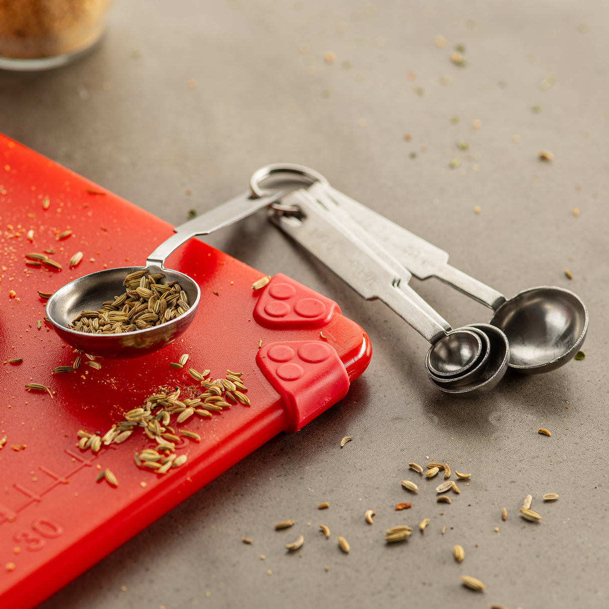 A set of stainless steel measuring spoons rests on a gray countertop, with one spoon holding fennel seeds. Seeds are scattered around, and the dishwasher safe spoons partially rest on a red silicone mat with a paw-shaped corner grip.