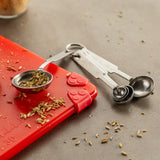 A set of stainless steel measuring spoons rests on a gray countertop, with one spoon holding fennel seeds. Seeds are scattered around, and the dishwasher safe spoons partially rest on a red silicone mat with a paw-shaped corner grip.