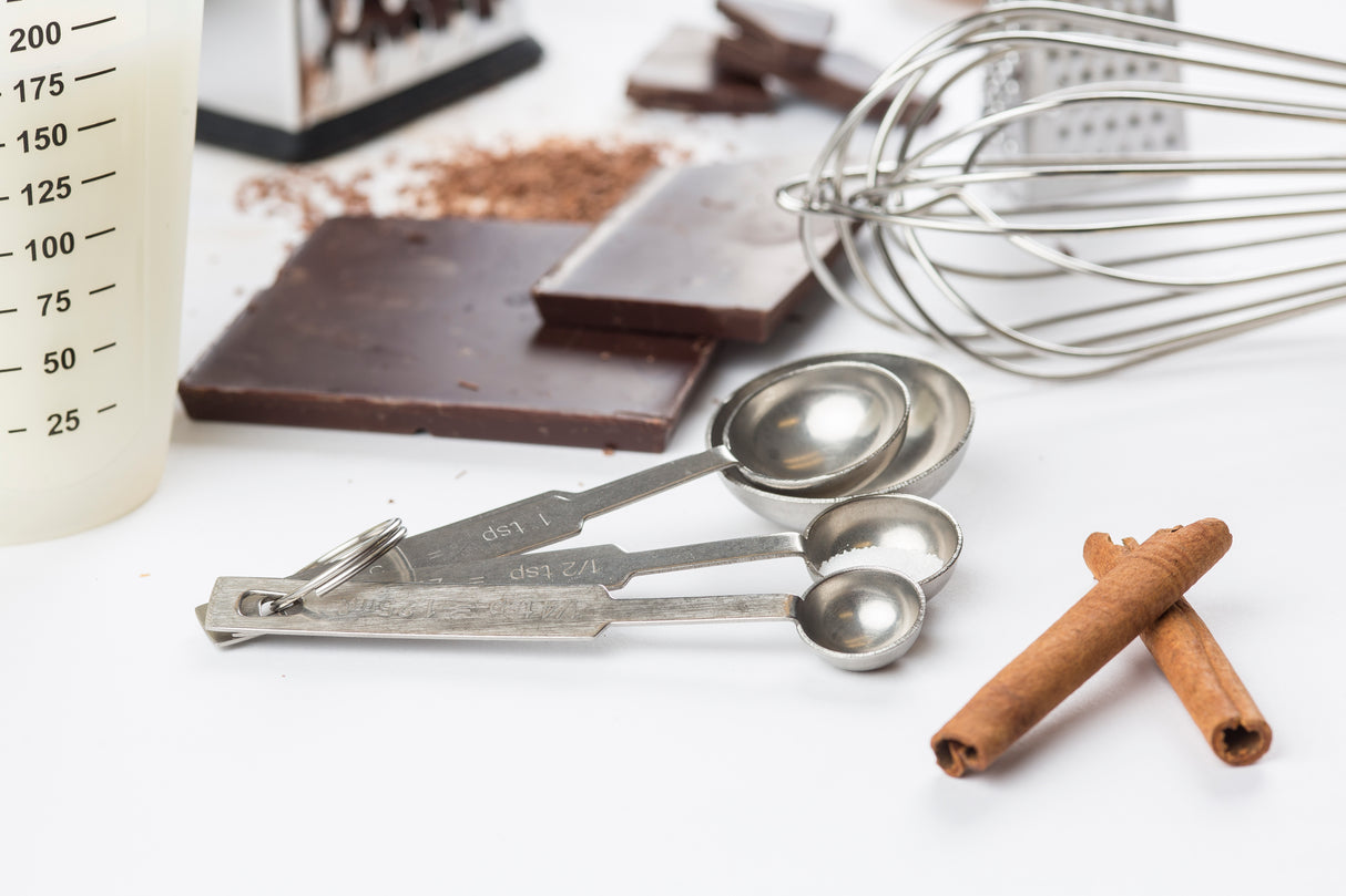 A set of stainless steel measuring spoons, two cinnamon sticks, chocolate pieces, a whisk, grated chocolate, and part of a measuring cup are arranged on a white surface, suggesting ingredients and tools for baking or cooking.