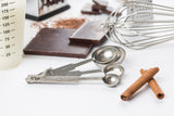 A set of stainless steel measuring spoons, two cinnamon sticks, chocolate pieces, a whisk, grated chocolate, and part of a measuring cup are arranged on a white surface, suggesting ingredients and tools for baking or cooking.