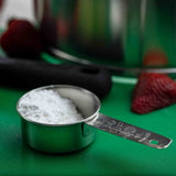 A stainless steel Measuring Cup filled with powdered sugar sits on a green surface. Blurry strawberries, a black utensil, and a stainless steel bowl appear in the background. The 1/4 cup measurement is marked on the cup’s handle.