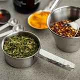 A stainless steel 1/4 cup is filled with dried green herbs. Nearby, another Measuring Cup holds red chili flakes and a spoon, while a third contains yellow turmeric powder. Measuring spoons rest on the softly blurred gray countertop.