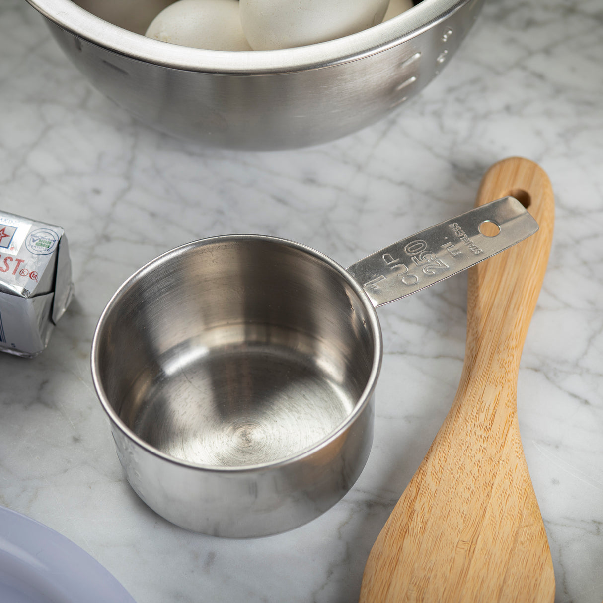 A stainless steel measuring cup marked “1/2 cup” sits on a marble countertop beside a wooden spoon, a stick of butter, and a metal bowl filled with white eggs, suggesting kitchen prep work or baking. This measuring cup is also dishwasher safe.