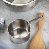 A stainless steel measuring cup marked “1/2 cup” sits on a marble countertop beside a wooden spoon, a stick of butter, and a metal bowl filled with white eggs, suggesting kitchen prep work or baking. This measuring cup is also dishwasher safe.