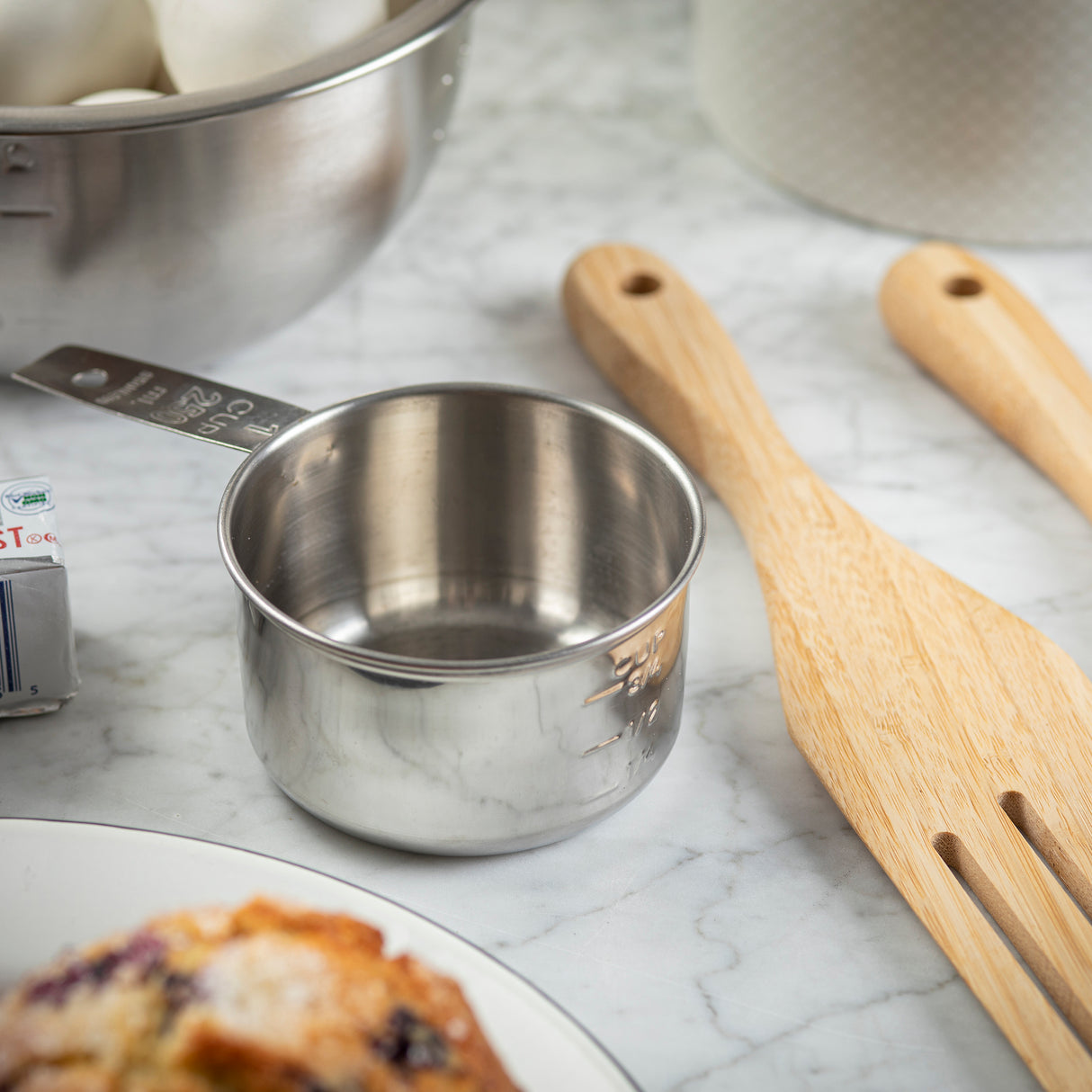 A stainless steel measuring cup sits on a marble countertop next to two light wooden utensils. In the background, there’s a metal bowl with white ingredients and a white container. A corner of foil-wrapped butter and a pastry on a plate are also partially visible.