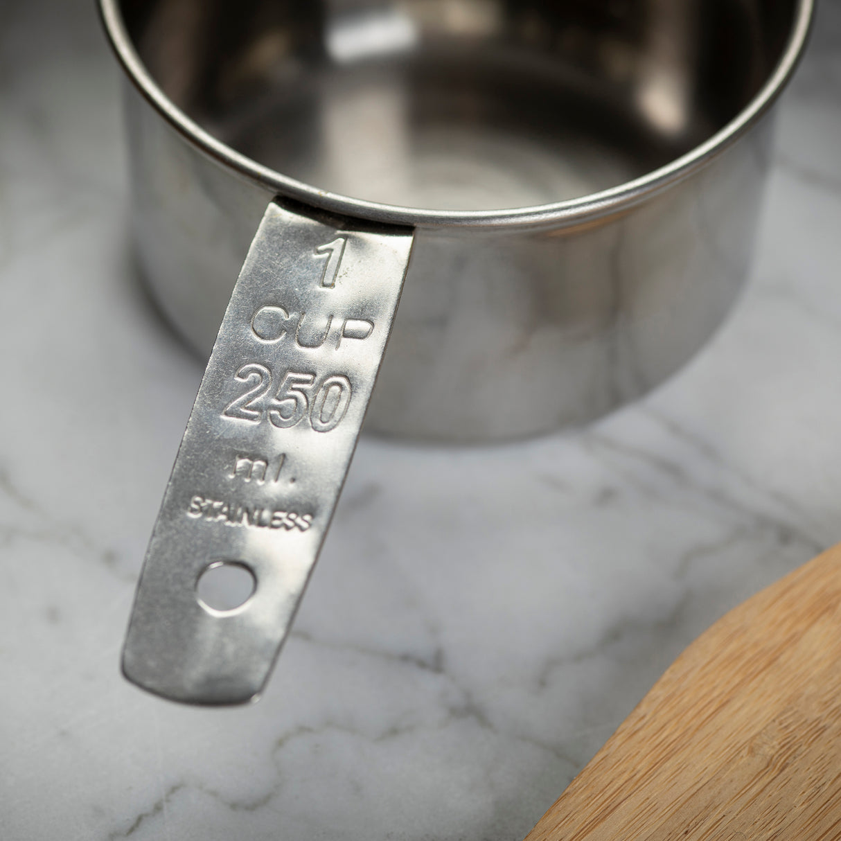 A close-up of a stainless steel measuring cup labeled “1 CUP 250 mL STAINLESS” sits on a marble countertop. The dishwasher safe handle with engraved measurements is in focus, while part of a wooden cutting board appears in the bottom right corner.