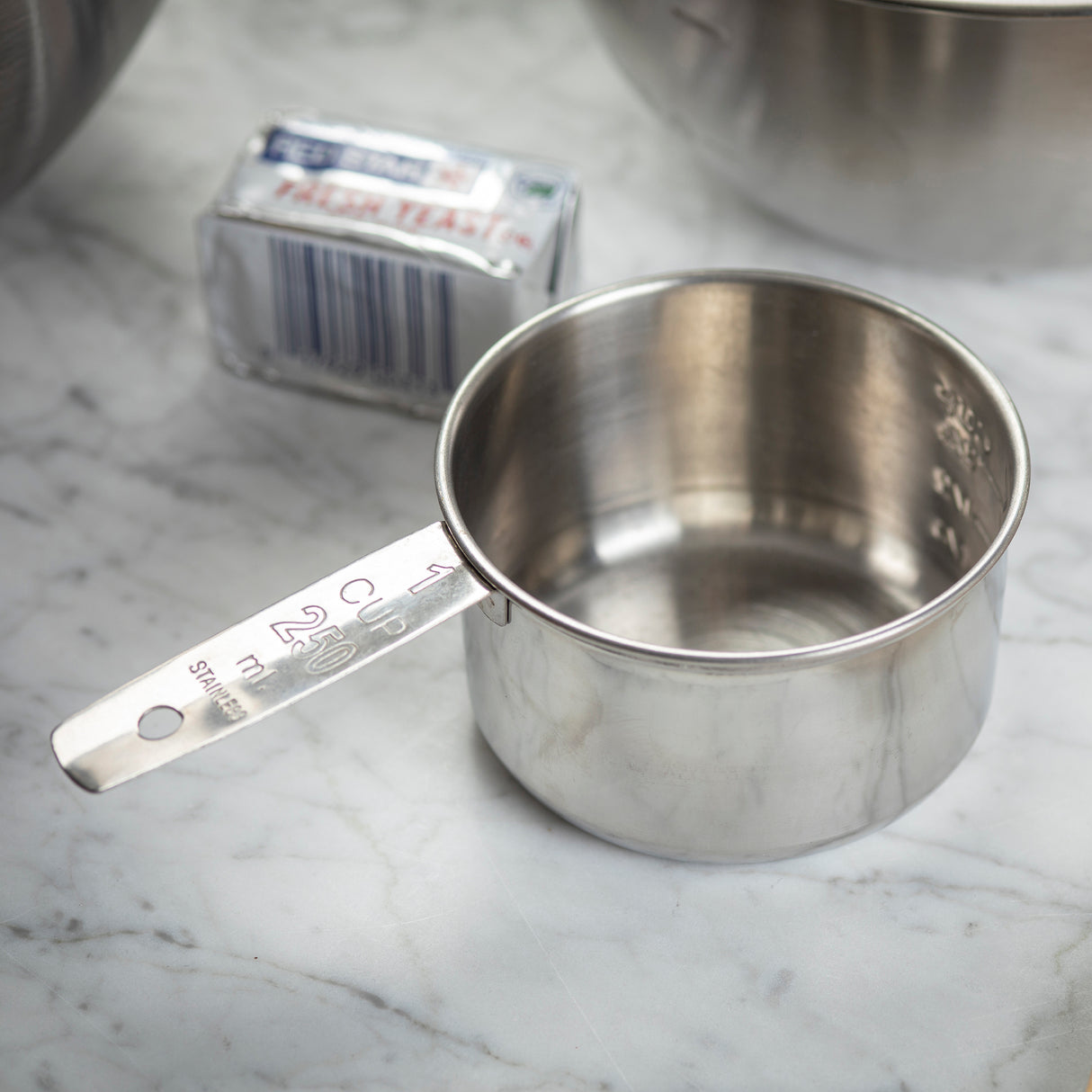 A stainless steel measuring cup marked “1 CUP 250 ml” sits on a marble countertop. Behind it is a wrapped stick of unsalted butter and part of a large metal mixing bowl, setting the scene for baking preparation. The cup is dishwasher safe.