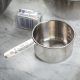 A stainless steel measuring cup marked “1 CUP 250 ml” sits on a marble countertop. Behind it is a wrapped stick of unsalted butter and part of a large metal mixing bowl, setting the scene for baking preparation. The cup is dishwasher safe.