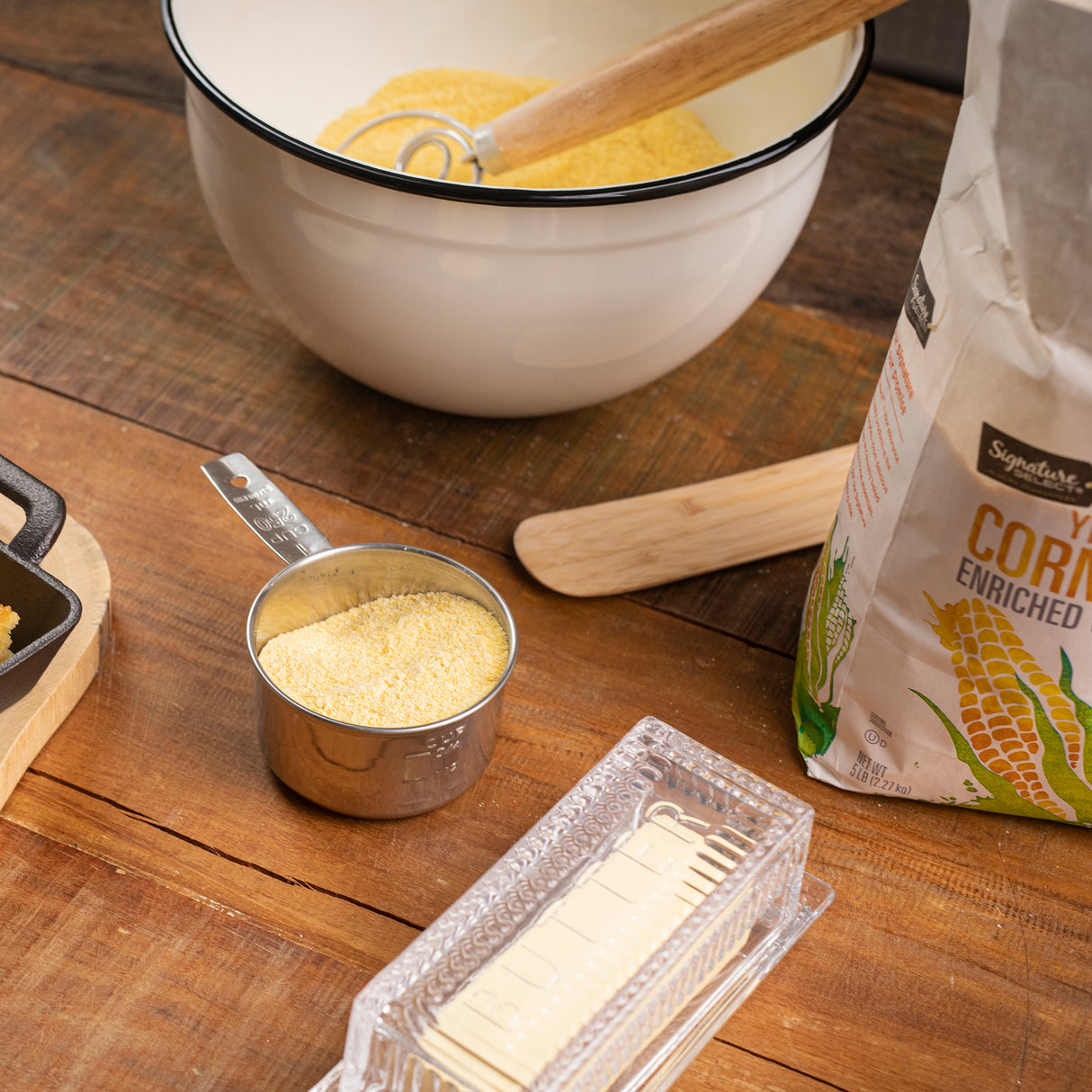 A wooden table holds a measuring cup filled with cornmeal, a stick of butter in a clear dish, a wooden spoon, an open bag of yellow cornmeal, and a dishwasher safe stainless steel mixing bowl with a whisk blending ingredients. Cornbread batter sits in a pan to the left.
