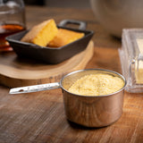 A stainless steel measuring cup filled with cornmeal sits on a wooden table. In the background, a small cast iron dish with cornbread, a stick of butter in a butter dish, and a glass with brown liquid are all slightly out of focus.