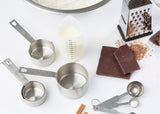 A flat lay of baking tools and ingredients: three stainless steel measuring cups, nested metal measuring spoons, a clear dishwasher safe measuring cup with liquid, dark chocolate bars, a box grater with chocolate shavings, cinnamon sticks, and flour—all on a white surface.