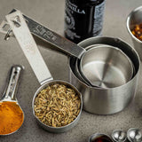 A set of stainless steel measuring cups and spoons rests on a gray surface. The smallest measuring cup holds fennel seeds, while a spoon displays vibrant turmeric powder. These dishwasher safe utensils add convenience to your kitchen.