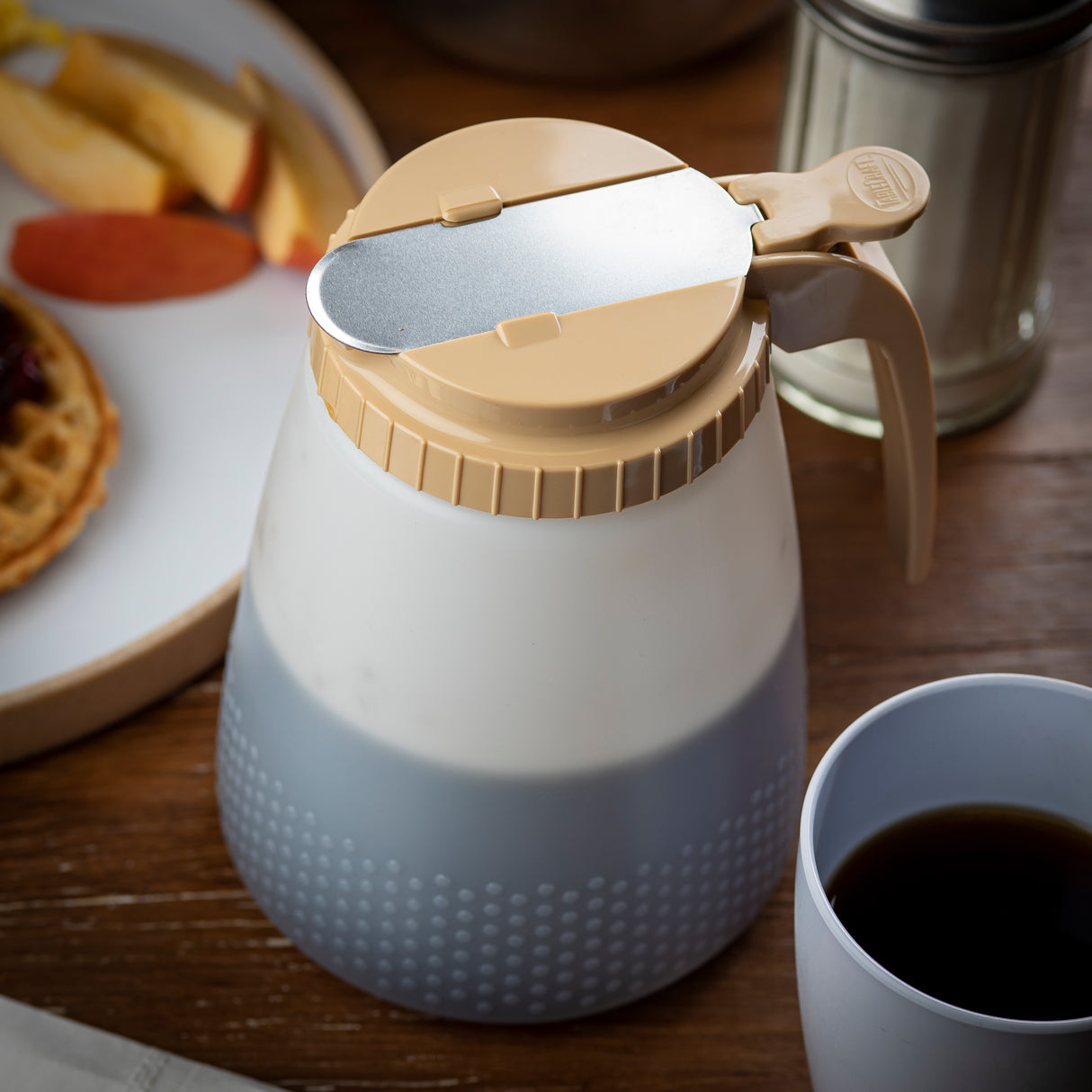 A BPA free All Purpose Dispenser in beige and white with a metal lid sits on a wooden table. Nearby are a white plate with waffles and apple slices, a metal shaker, and a mug of dark coffee, creating a cozy breakfast scene.