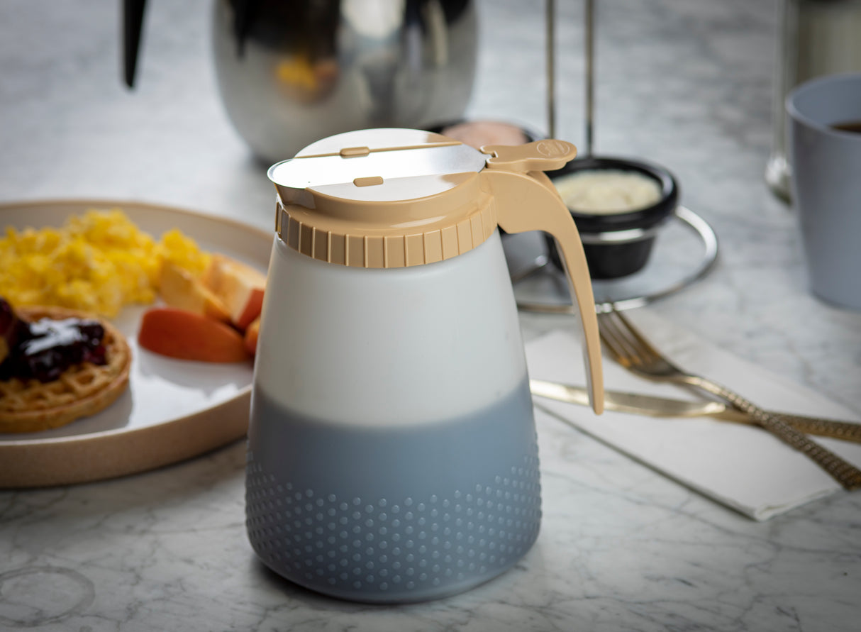 A BPA free, all purpose dispenser with a beige lid sits on a marble table. In the background, there’s a plate with scrambled eggs, sausage, and a waffle with jam—perfect for a cozy breakfast setting.