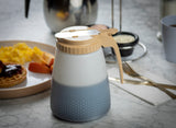 A BPA free, all purpose dispenser with a beige lid sits on a marble table. In the background, there’s a plate with scrambled eggs, sausage, and a waffle with jam—perfect for a cozy breakfast setting.