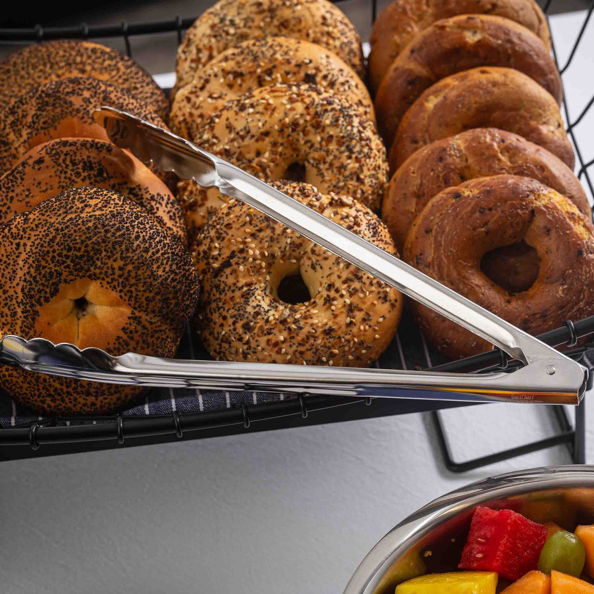 A tray of assorted bagels—including everything, poppy seed, and plain—sits on a wire rack. Dishwasher safe stainless steel utility tongs rest on the tray. In the bottom right corner, a bowl of colorful cut fruit is partially visible.