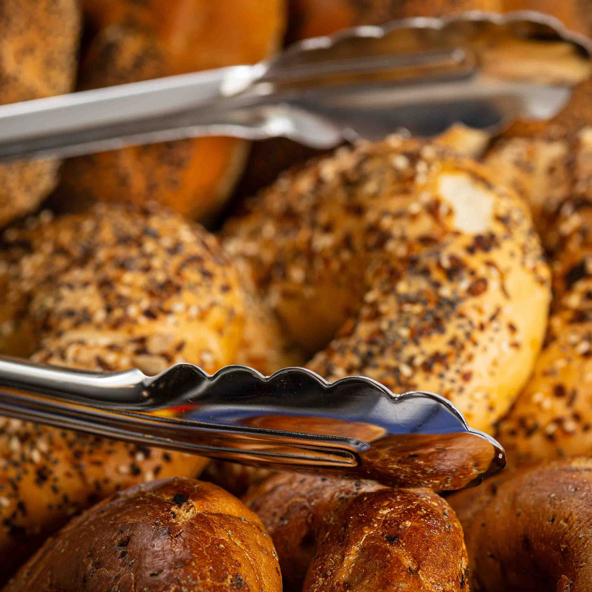 A close-up of stainless steel Utility Tongs gripping an everything bagel, surrounded by golden brown bagels with various toppings. The tongs are dishwasher safe, and the image radiates freshness and warm, inviting tones.