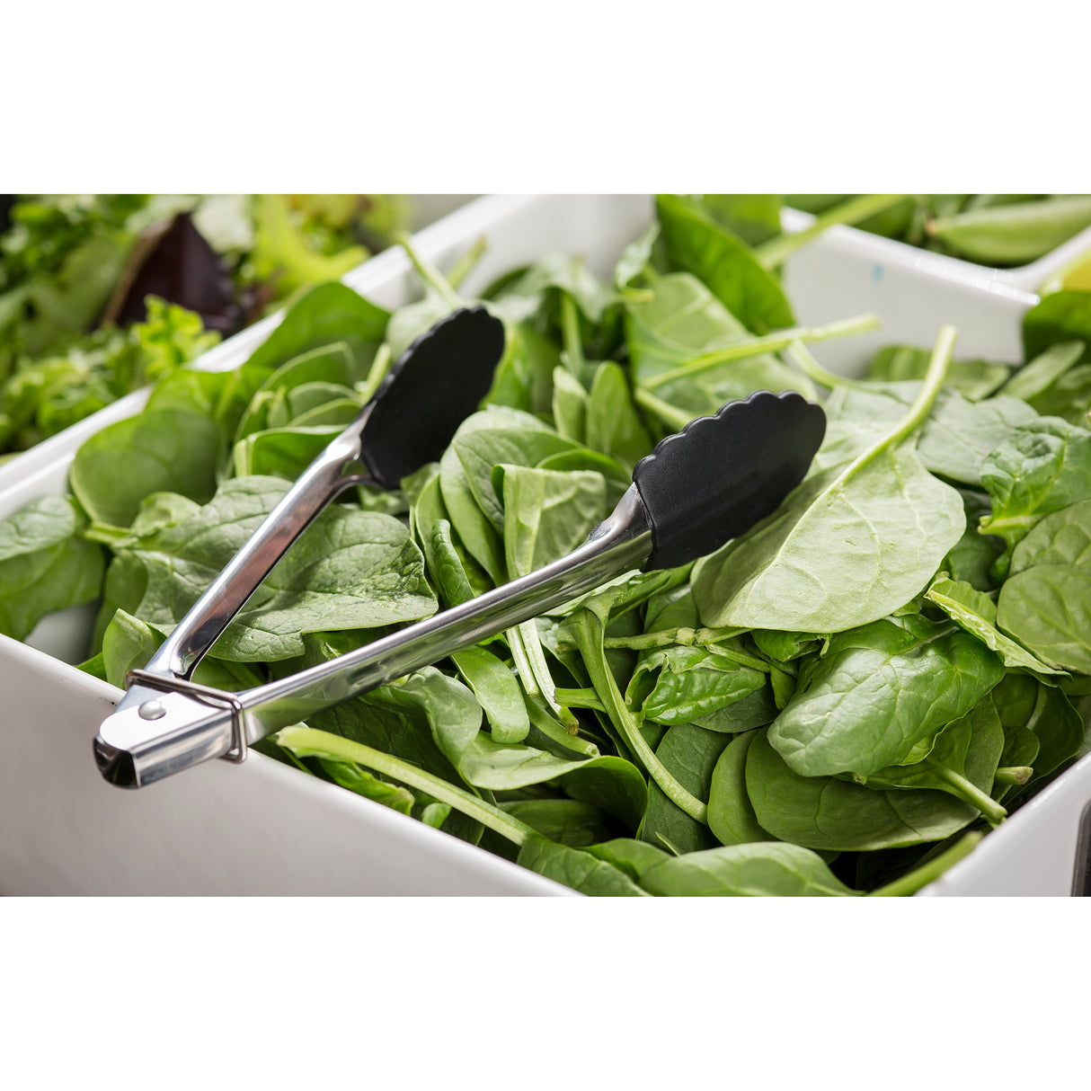 A pair of stainless steel utility tongs with black silicone tips rests on a pile of fresh spinach leaves in a white rectangular container, likely at a salad bar or buffet. They're durable and dishwasher safe for easy cleaning. Other leafy greens are nearby.