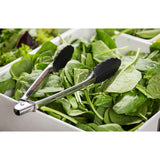 A pair of stainless steel utility tongs with black silicone tips rests on a pile of fresh spinach leaves in a white rectangular container, likely at a salad bar or buffet. They're durable and dishwasher safe for easy cleaning. Other leafy greens are nearby.