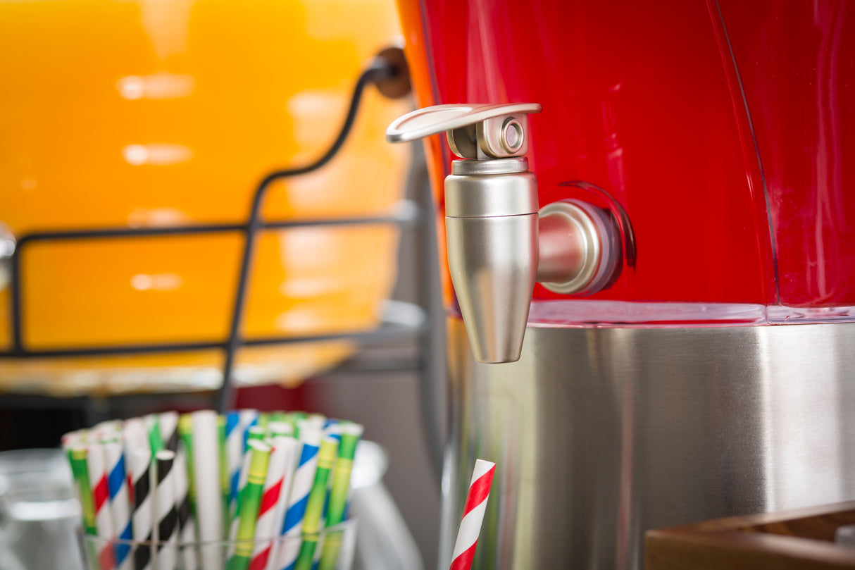 Close-up of a drink dispenser featuring a nickel plated beverage dispenser faucet on a red container. In the background, there's an orange dispenser and colorful striped paper straws in a holder at the lower left corner.