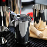 A shiny stainless steel Bar Shaker with a strainer and bar spoon rests on a black mat. Nearby are a jigger, muddler on a wooden board, and extra shaker tins. The dishwasher safe pieces reflect light and surrounding bar tools beautifully.