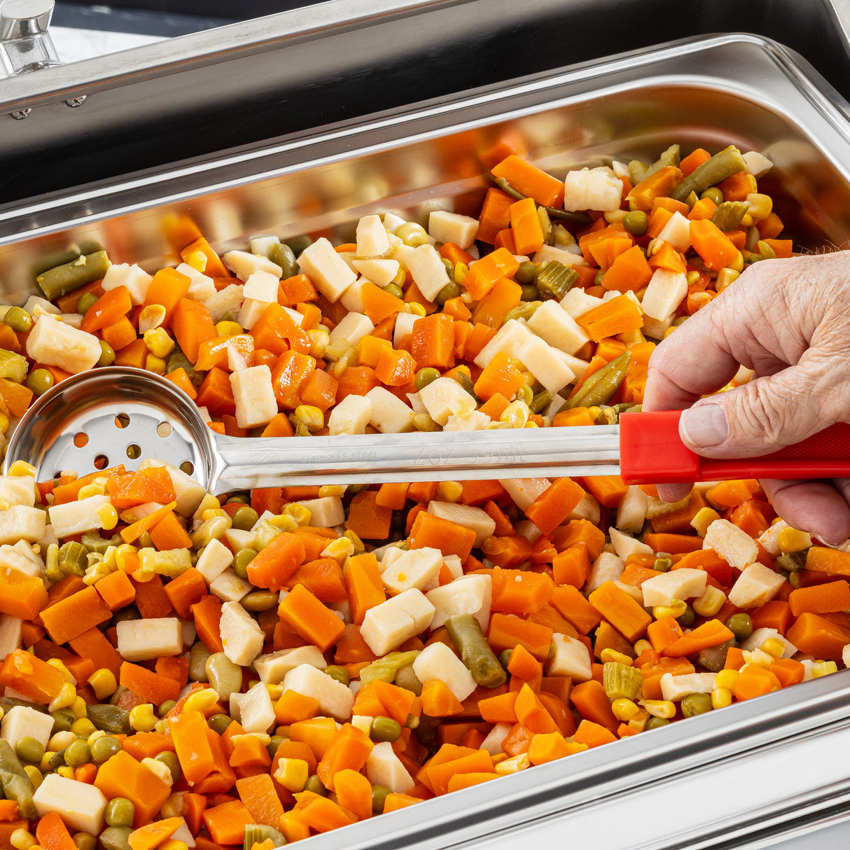 A close-up of a metal pan filled with diced mixed vegetables—carrots, green beans, corn, and potatoes. A hand uses a perforated stainless steel serving spoonout with a red handle to scoop the vegetables at a buffet or cafeteria setup.