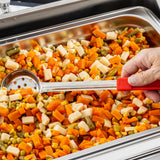 A close-up of a metal pan filled with diced mixed vegetables—carrots, green beans, corn, and potatoes. A hand uses a perforated stainless steel serving spoonout with a red handle to scoop the vegetables at a buffet or cafeteria setup.
