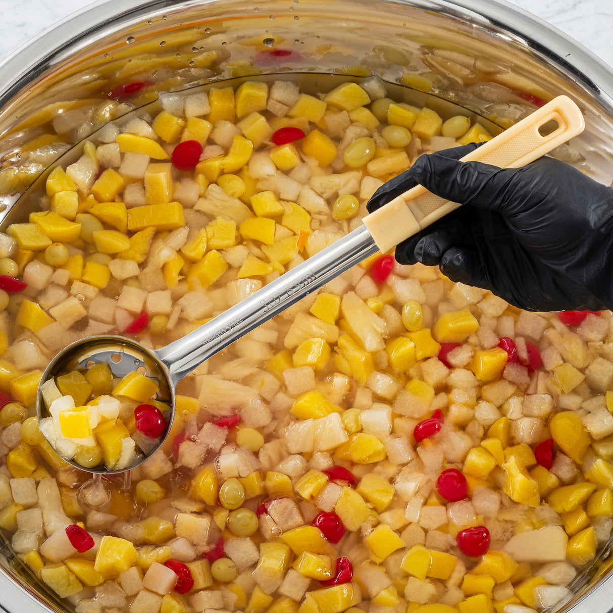 A gloved hand holds a stainless steel perforated ladle, spooning out colorful fruit cocktail from a large bowl filled with diced peaches, pears, pineapple, grapes, and cherries in syrupy liquid. The ladle displays a vibrant mix of fruit pieces over the bowl.