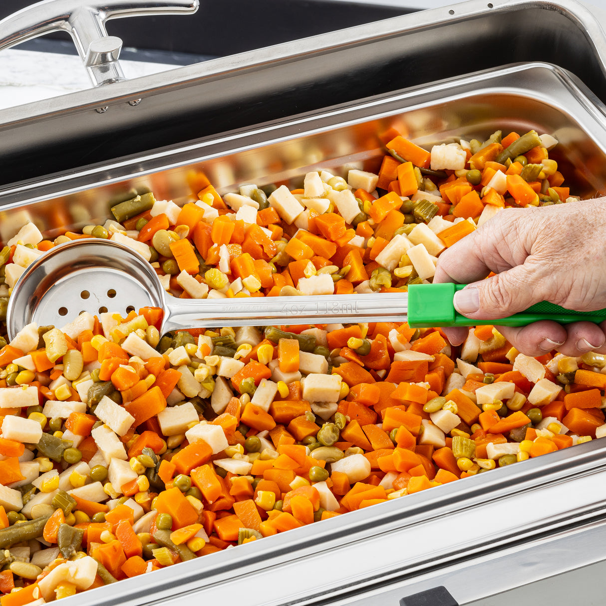 A hand holding a green-handled perforated Spoonout scoops a mix of diced cooked vegetables—including carrots, green beans, corn, and parsnips—from a stainless steel chafing dish.