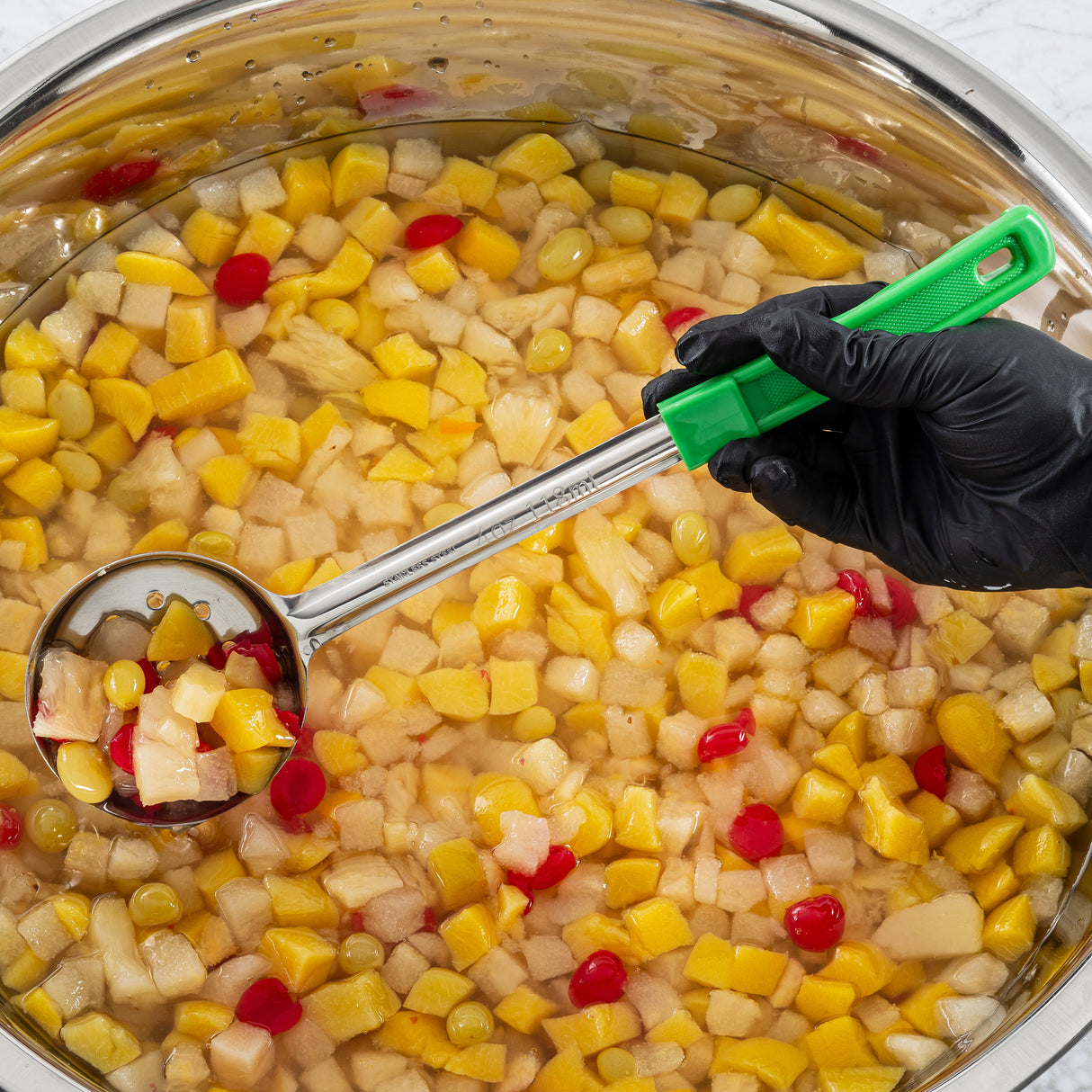 A gloved hand holds a perforated Spoonout ladle scooping fruit cocktail from a large stainless steel pot filled with diced peaches, pears, grapes, pineapple, and red cherries in syrup. The green-handled ladle lets the colorful fruit drain evenly.
