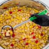 A gloved hand holds a perforated Spoonout ladle scooping fruit cocktail from a large stainless steel pot filled with diced peaches, pears, grapes, pineapple, and red cherries in syrup. The green-handled ladle lets the colorful fruit drain evenly.