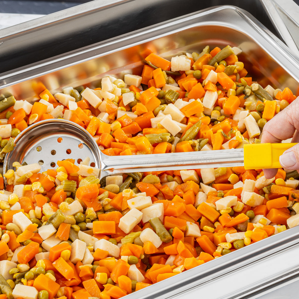 A hand holds a yellow-handled perforated Spoonout above a stainless steel tray filled with a colorful mix of steamed diced vegetables, including carrots, green beans, corn, and potatoes, ready to be served.