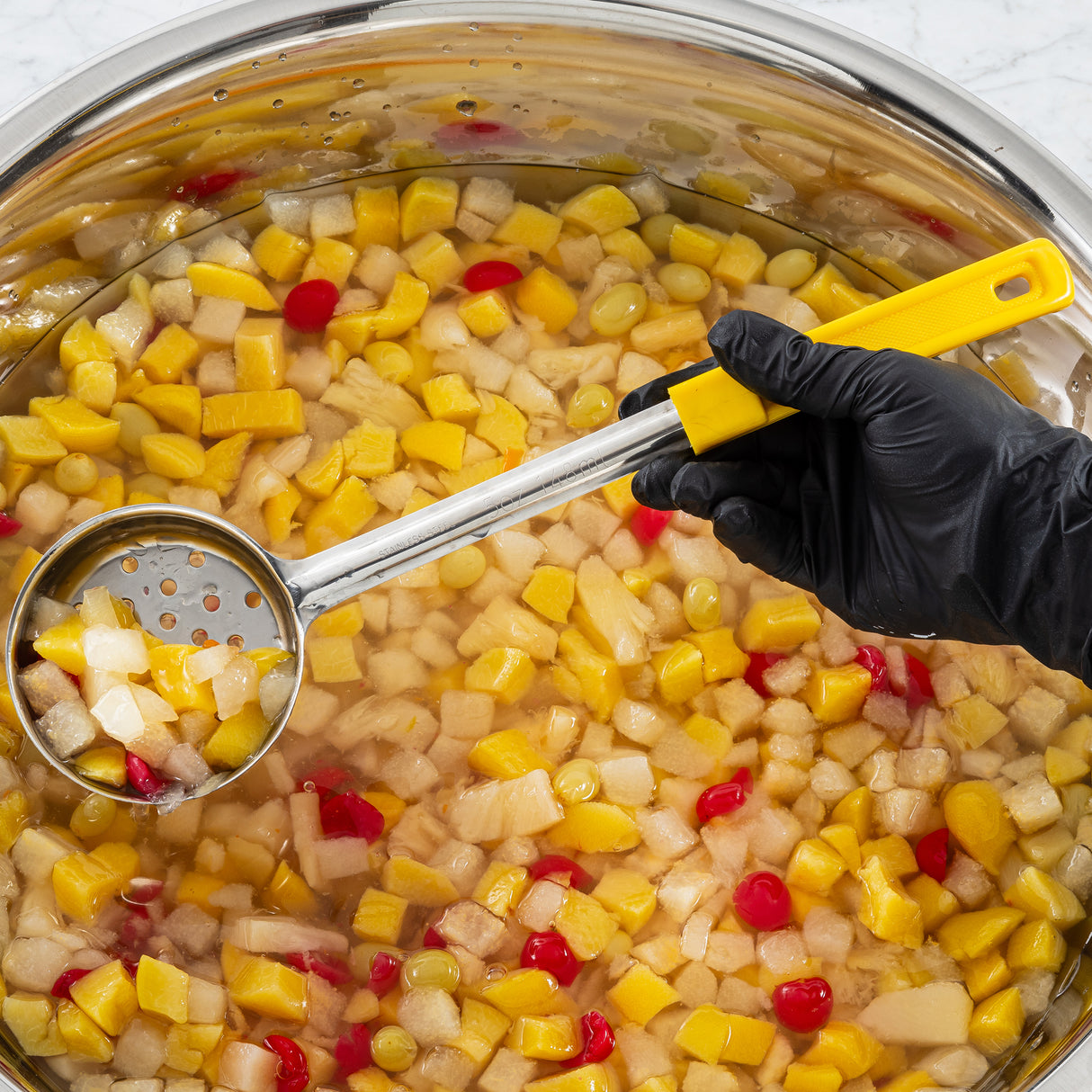 A hand in a black glove uses a perforated spoon with a yellow handle to scoop up pineapple, pear, peach, and red cherries from a stainless steel bowl filled with clear syrup. The brightly colored fruit floats invitingly in the liquid.