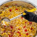 A hand in a black glove uses a perforated spoon with a yellow handle to scoop up pineapple, pear, peach, and red cherries from a stainless steel bowl filled with clear syrup. The brightly colored fruit floats invitingly in the liquid.