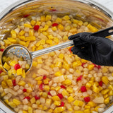 A gloved hand holds a perforated stainless steel Spoonout spoon over a large metal pot filled with canned fruit cocktail in syrup—diced yellow peaches, pears, grapes, and red cherries—ready to be served or prepared further.