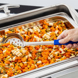 A close-up of a hand holding the Spoonout slotted serving spoon with a blue handle, scooping a colorful mix of diced veggies from a stainless steel food warmer tray; the Spoonout is durable and dishwasher safe.