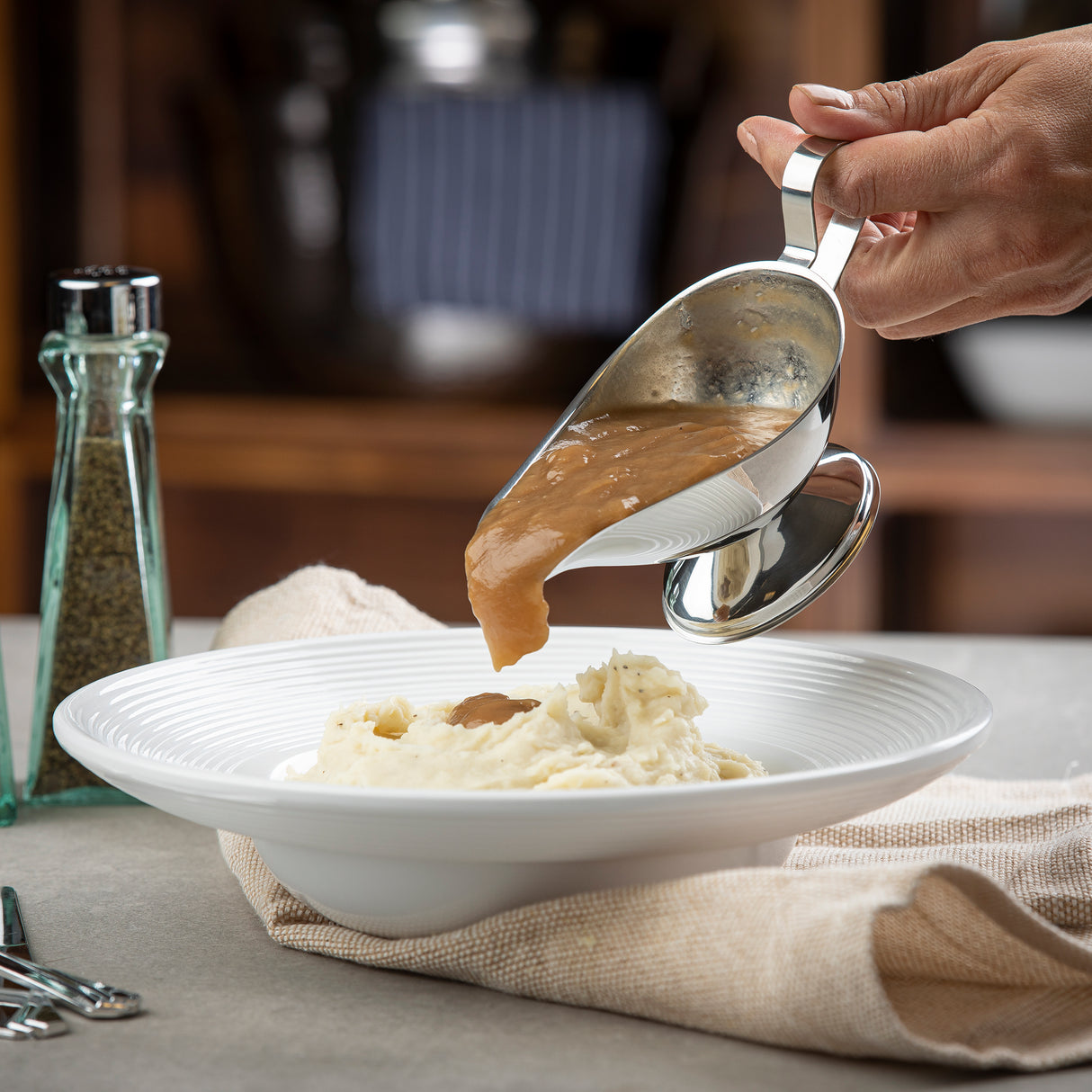 A hand pours brown gravy from an 18/8 stainless steel Gravy Boat onto a mound of mashed potatoes in a white bowl. A pepper shaker and beige napkin sit nearby on the table, with a blurred kitchen background. The Gravy Boat is dishwasher safe.
