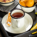 A white porcelain enamel mug with a dark rim sits on a matching plate, filled with tea. Next to the mug are two orange sugar packets. Nearby are a bowl of oranges, granola, and part of a banana, all on a black tray.