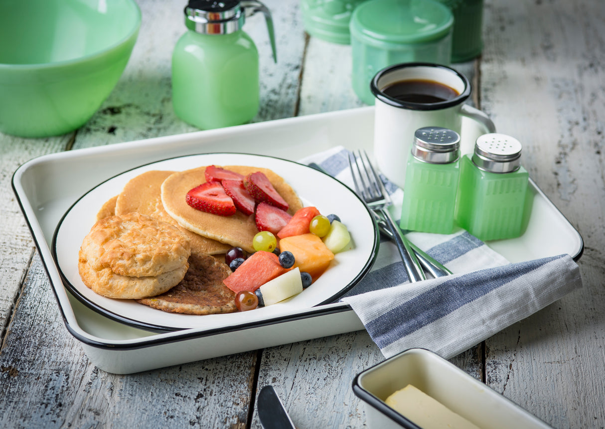 A porcelain enamel coated steel Enamelware serving tray holds a plate with pancakes topped with strawberries, a biscuit, sausage patty, and fruit. Nearby are dishwasher safe green glass syrup dispensers, coffee, utensils, and a butter dish on a rustic white table.