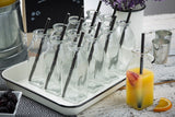 A tray of empty glass milk bottles with black straws sits on a rustic white table. Nearby, a bottle filled with orange juice, fruit slices, and a slice of orange rest on an Enamelware Serving Tray, with blackberries and lavender flowers in the background.